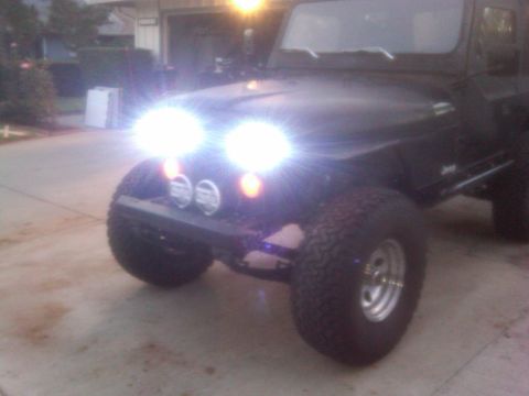 1986 Ford Bronco - A black Jeep Wrangler is shown from the front, its bright headlights illuminating the scene. The vehicle has off-road tires and appears to…