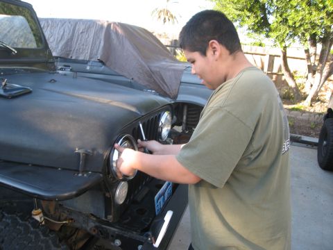 1986 Ford Bronco - A person works on the headlight of a black Jeep CJ, with another vehicle partially visible behind it.
