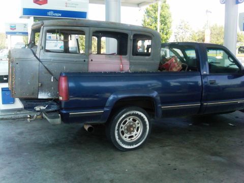 1986 Ford Bronco - A vintage car body is transported in the bed of a blue pickup truck, with gas station signage visible in the background.