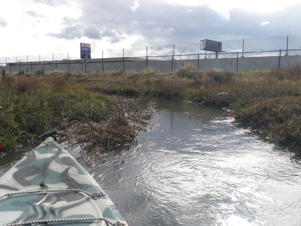 Kayak moving through a narrow waterway with overgrown banks. A chain fence and billboards are visible in the background