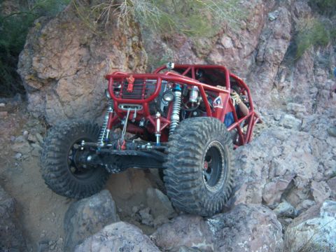 Suzuki Samurai - A red tube-chassis rock buggy climbing a boulder-strewn desert trail with its front axle flexed and the driver's arm out of the cage. Coilo…