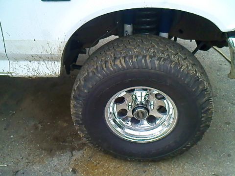 1995 Ford F-150 - Close-up of a lifted white truck's front wheel and fender, showing off-road tires and suspension components.