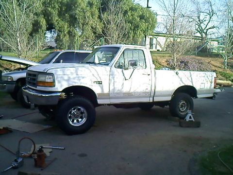 1995 Ford F-150 - A white 1995 Ford F-150 pickup truck is shown in profile, a lifted suspension and another vehicle in the background.