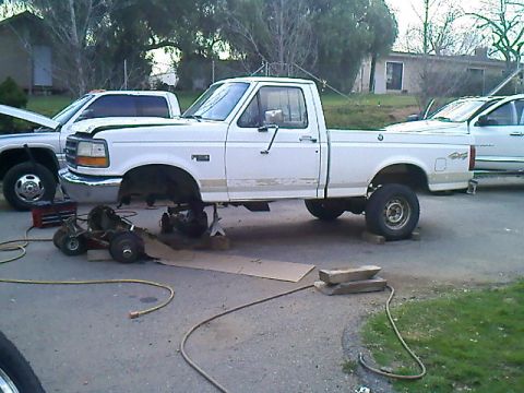 1995 Ford F-150 - A white pickup truck is elevated on jack stands, with its front end undergoing maintenance. Other vehicles are parked in the background.