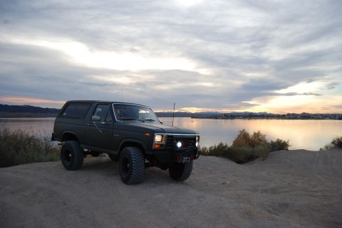 1982 Ford Bronco - A lifted Ford Bronco sits on a dusty overlook as the sun sets over a calm body of water.