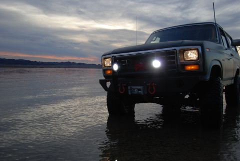 1982 Ford Bronco - Ford Bronco sits in shallow water as the sun sets, casting reflections on the wet surface.