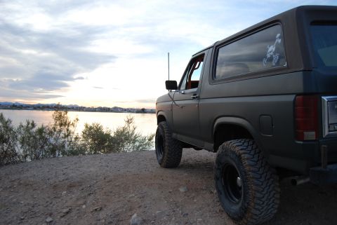 1982 Ford Bronco - A dark green Ford Bronco sits by a lake at sunset, its lifted suspension and large tires hinting at off-road capabilities.