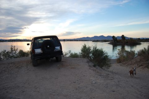 1982 Ford Bronco - A black SUV is parked on a dirt embankment overlooking a lake as the sun sets, with a dog running in the foreground.