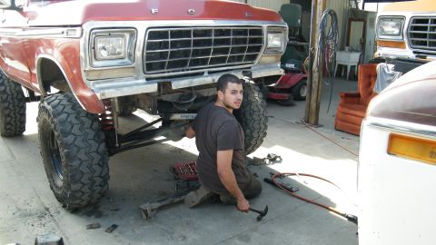1982 Ford Bronco - Mechanic works on the front end of a lifted 1979 Ford truck in a garage. Another vehicle is parked nearby.
