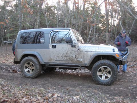 2005 Jeep Wrangler - A mud-splattered 2005 Jeep Wrangler sits on a trail surrounded by trees, with another vehicle in the background.