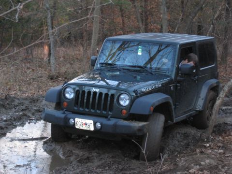2005 Jeep Wrangler - A dark green Jeep Wrangler Rubicon navigates a muddy trail, its tires kicking up dirt and water.