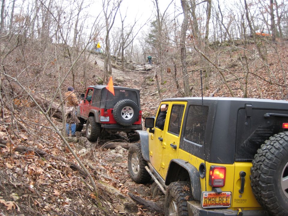 Yellow Jeep Wrangler and red Jeep Wrangler driving up a rocky, wooded trail. The yellow Jeep is in the foreground