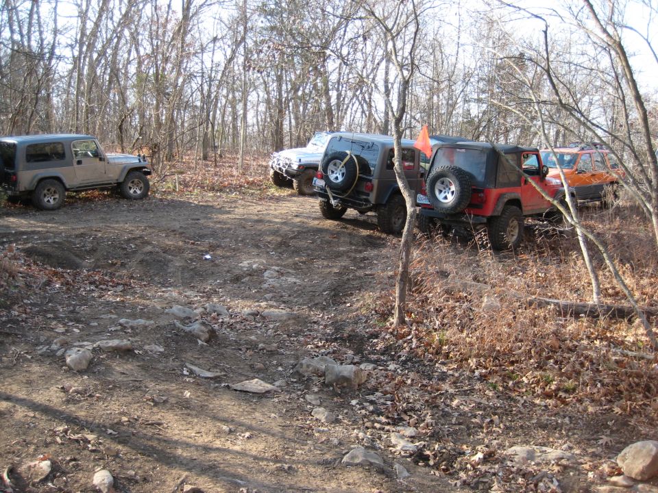 Four Jeep Wranglers parked on a dirt path in a wooded area; one is tan, one is dark gray, one is red, and one is orange