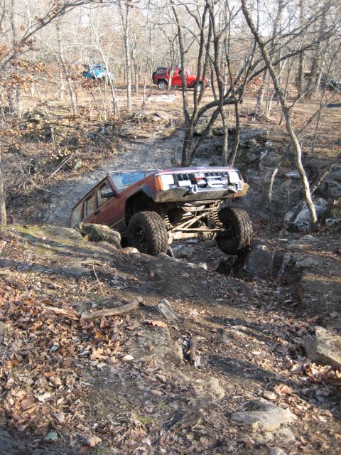 2005 Jeep Wrangler - A lifted maroon Jeep Cherokee XJ crawls nose-up over a rock ledge on a leaf-covered winter trail, flexing its front suspension with one whe…