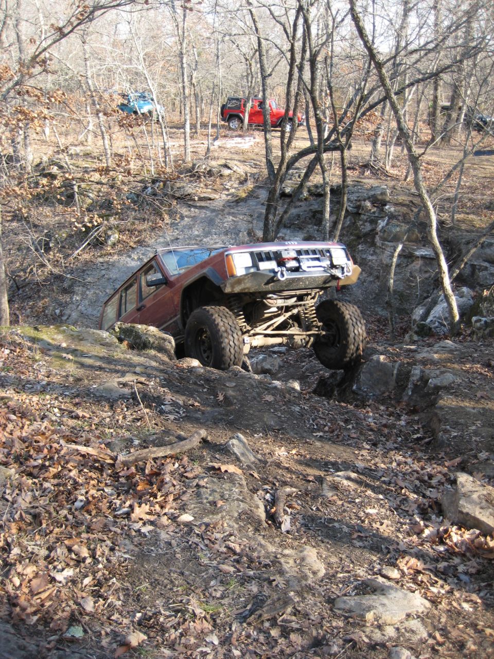 Lifted maroon Jeep Cherokee XJ climbing a rock ledge on a wooded winter trail