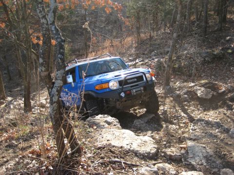 2005 Jeep Wrangler - A blue Toyota FJ Cruiser tackles a challenging off-road trail with large rocks and trees.