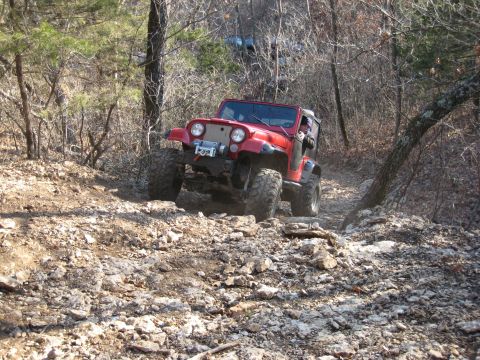 2005 Jeep Wrangler - A red Jeep Wrangler tackles a challenging, rocky trail through a wooded area. Another vehicle can be seen further up the path.