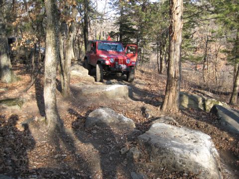 2005 Jeep Wrangler - A red Jeep Wrangler navigates a challenging rocky trail through a wooded area, with another vehicle partially visible in the background.