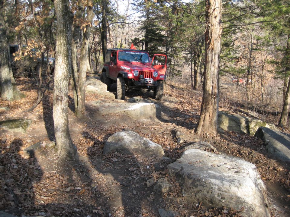 Red Jeep Wrangler TJ on a rocky trail, front passenger door open. Text on windshield reads 'Ruger's Ride'