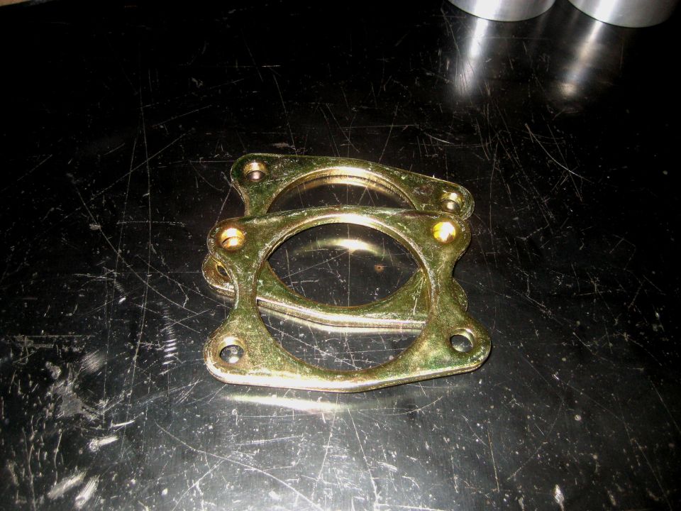 Two gold-colored exhaust gaskets with four bolt holes each, stacked on a dark, scratched surface