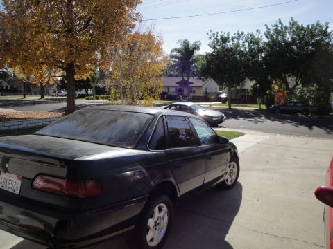 1995 Ford Taurus - This dark sedan, identified as a 1995 Ford Taurus, is parked in a driveway with other vehicles visible in the background.