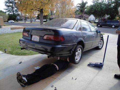 1995 Ford Taurus - A person works on the exhaust of a dark blue 1995 Ford Taurus, lifted by a jack.
