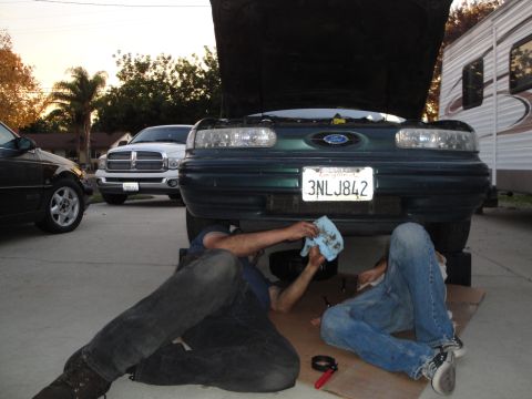 1995 Ford Taurus - Two individuals attend to maintenance on a 1995 Ford Taurus, its hood raised to reveal the engine bay.