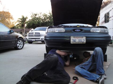 1995 Ford Taurus - Two mechanics work on the underside of a 1995 Ford Taurus, with other vehicles nearby.