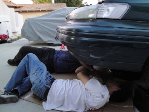 1995 Ford Taurus - Two individuals are engaged in maintenance work beneath a vehicle, with another car partially visible in the background.