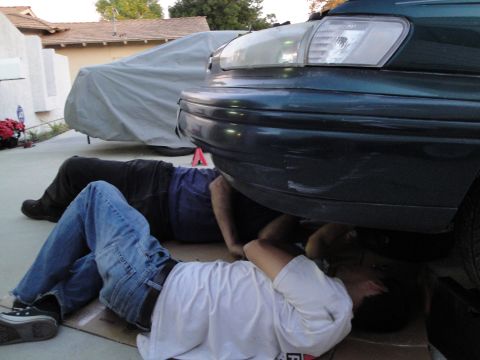 1995 Ford Taurus - Two individuals are working on the underside of a dark green 1995 Ford Taurus.