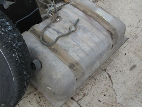 1986 Ford Bronco - A metal gas tank with attached fuel lines and securing straps is shown next to a tire.
