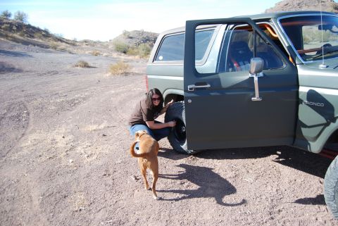 1982 Ford Bronco - A woman changes a tire on a 1982 Ford Bronco in a desert landscape.