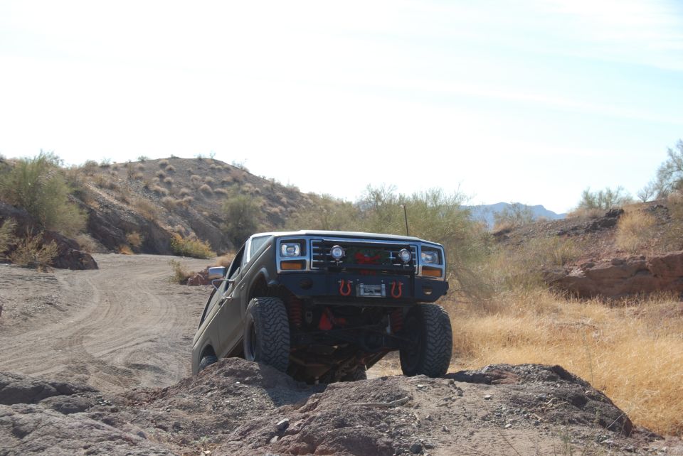 Dark green 1980s Ford Bronco off-roading on a rocky, desert trail with a modified bumper and large tires