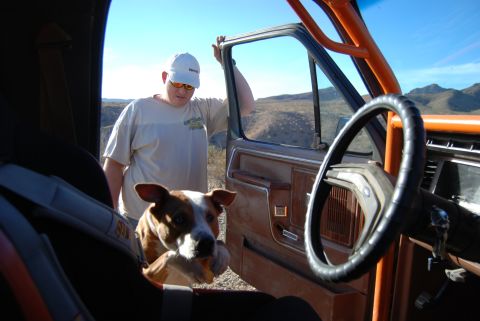 1982 Ford Bronco - Interior view of a 1982 Ford Bronco, a roll cage, a dog, and a man standing by the open door in a desert landscape.