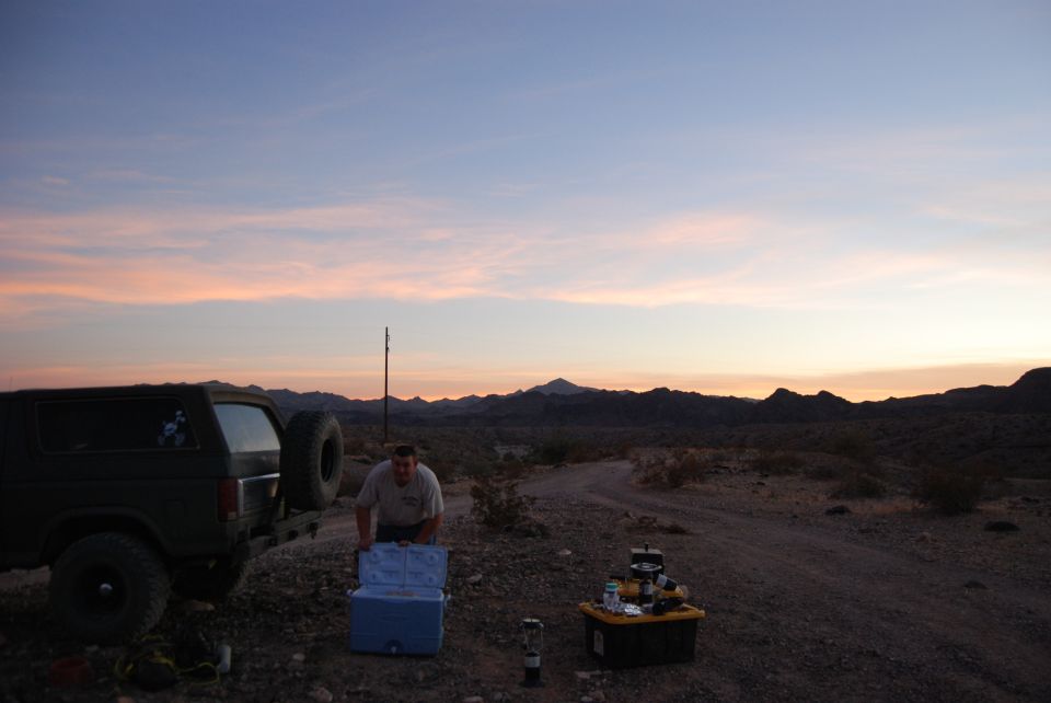 Man by cooler next to a dark-colored Ford Bronco with a spare tire on the back, desert landscape at dusk
