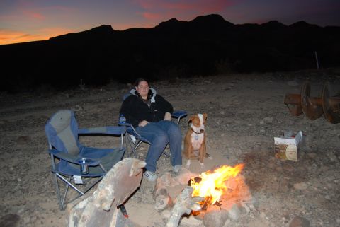 1982 Ford Bronco - A woman and her dog relax by a campfire as the sun sets behind a mountain range.