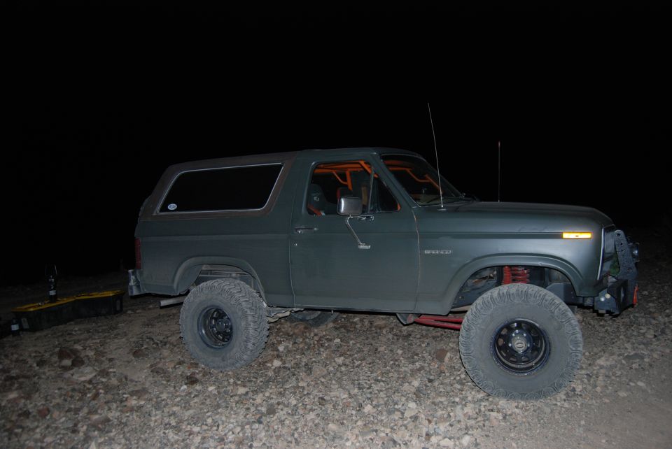 Dark green Ford Bronco with orange roll cage, lifted suspension, and large tires parked on gravel at night
