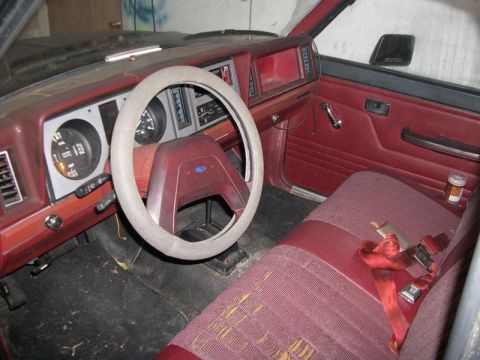 1987 Ford Ranger - The interior of a 1987 Ford Ranger shows its red fabric seats, cracked dashboard, and a Ford emblem on the steering wheel.