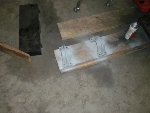 1995 Ford Bronco - Two metal brackets, likely automotive parts, are shown after being spray-painted and placed on a wooden plank on a concrete floor.