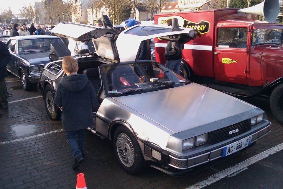 DeLorean DMC-12 with gullwing doors open, a boy walks past. Red truck in background