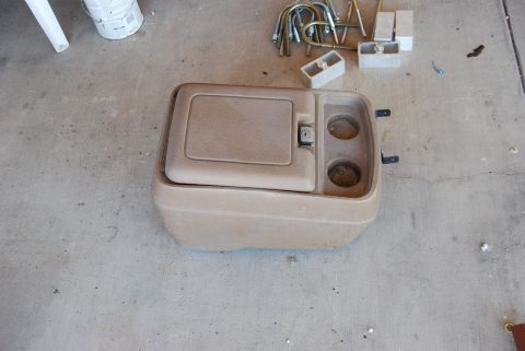 1982 Ford Bronco - A vehicle's center console, likely removed for repair or replacement, rests on a concrete surface with various hardware scattered nearby.