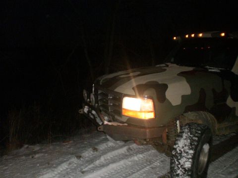 1995 Ford Bronco - A camouflaged Ford truck is seen in a snowy environment at night, illuminated by its headlights and roof lights.