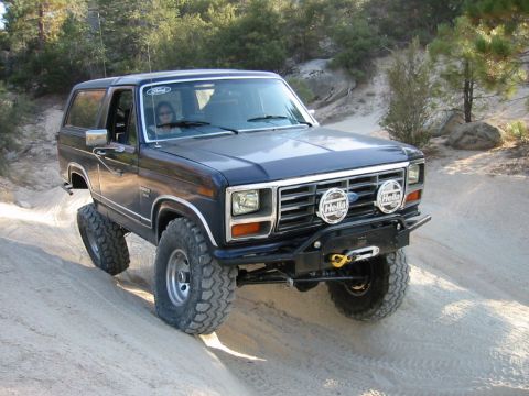 1985 Ford Bronco - A dark blue Ford Bronco navigates a sandy, off-road trail with trees and rocks in the background.
