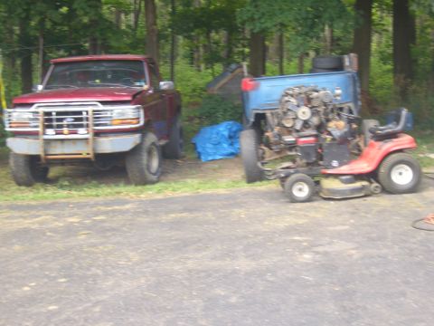 1992 Ford F-150 - A red pickup truck and a disassembled riding mower sit in a grassy area next to a paved surface.