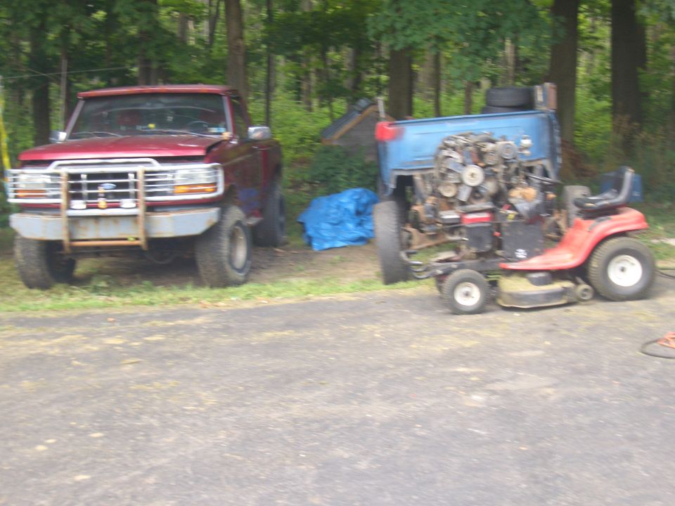 Red pickup truck with a large metal bumper and a blue tarp behind it. A red riding mower with its engine exposed is to the right