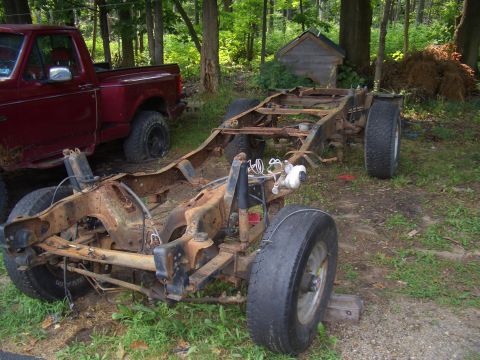 1992 Ford F-150 - Rusted truck chassis and frame sits in a yard next to a red truck.