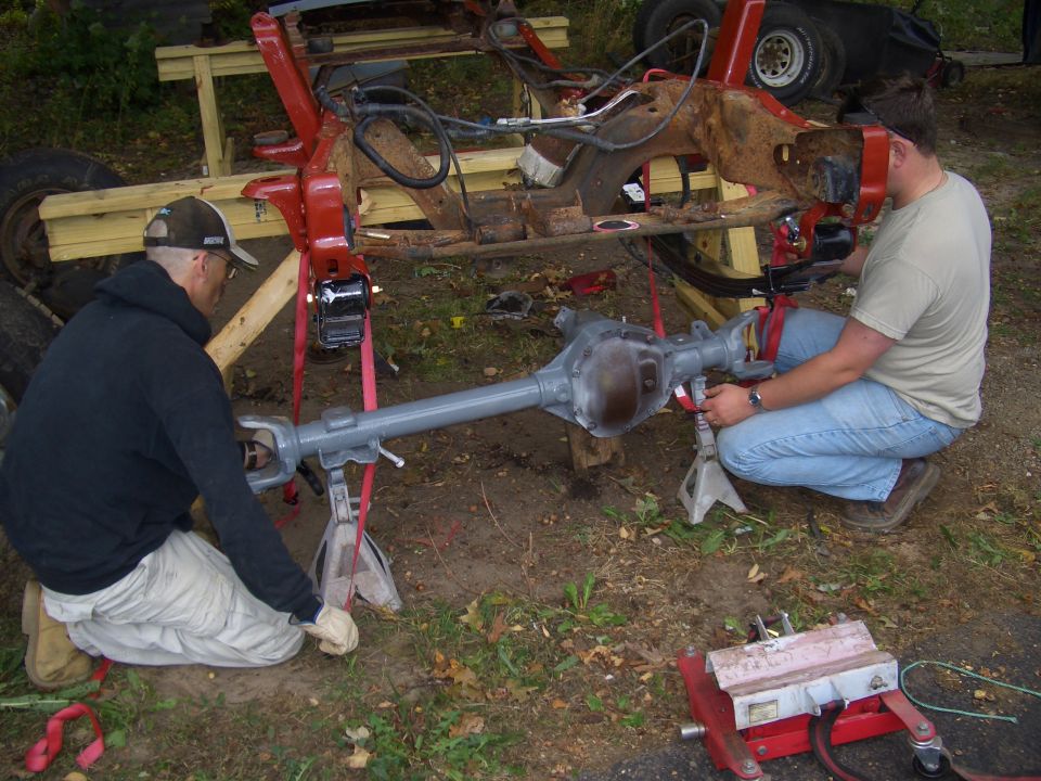 Two guys working on a red truck frame and gray rear axle assembly supported by jack stands