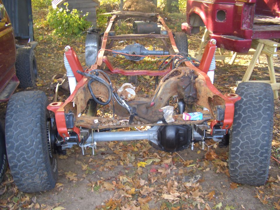 rusted frame of a truck with suspension and axles attached, next to a red vehicle body