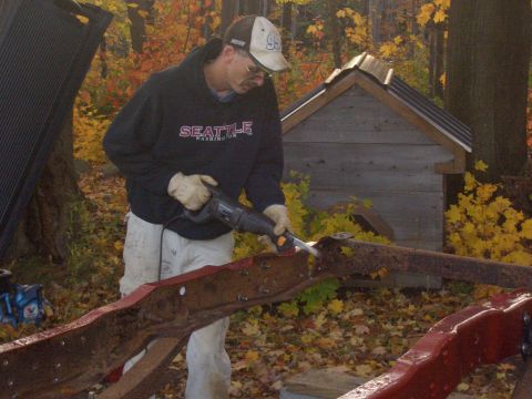 1992 Ford F-150 - A man uses a reciprocating saw to cut a heavily rusted truck frame in an outdoor setting with autumn foliage.