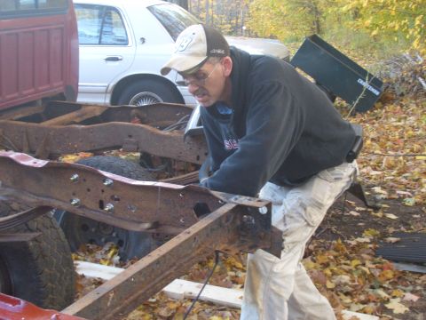 1992 Ford F-150 - A man works on the heavily rusted frame of a red truck, with a white car visible behind it.